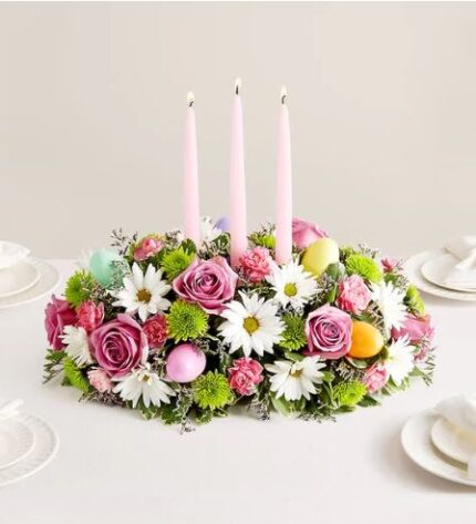 A lush Easter floral centerpiece featuring pink roses, white daisies, green chrysanthemums, and pink carnations, accented with three tall pink taper candles and colorful Easter eggs on a white tablecloth.