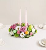 A colorful Easter floral centerpiece on a white dining table featuring pink roses, white daisies, green chrysanthemums, and two tall pink taper candles, accented with pastel-colored Easter eggs.
