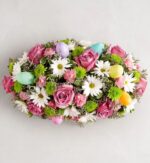 A lush, oval-shaped Easter floral centerpiece featuring pink roses, white daisies, and green chrysanthemums, decorated with pastel-colored Easter eggs in shades of purple, blue, yellow, and green.