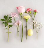 A flat-lay arrangement of various spring flowers including pink roses, carnations, daisies, baby's breath, a leafy green stem, and a single yellow Easter egg on a light gray background.