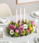 A festive Easter table centerpiece featuring pink roses, green chrysanthemums, white daisies, and pink carnations arranged with three tall pink taper candles and colorful Easter eggs on a grey tablecloth.
