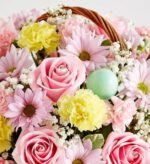 A close-up of a floral Easter basket featuring pink roses, yellow carnations, light purple daisies, baby's breath, and a single light green Easter egg nestled among the blooms.