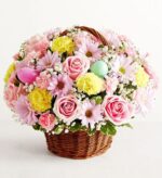 A lush Easter flower arrangement in a brown wicker basket featuring pink roses, yellow carnations, light purple daisies, and decorative Easter eggs.
