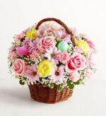 A woven brown wicker basket filled with a dense, rounded floral arrangement of pink roses, yellow carnations, light purple daisies, and white baby's breath, featuring decorative pink and light green Easter eggs tucked among the blooms.