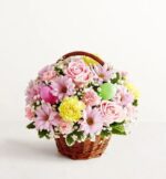 A small wicker basket filled with a dense arrangement of pastel-colored spring flowers, including pink roses, lavender daisies, yellow carnations, and small pink carnations, accented with bright pink and lime green Easter eggs and white baby's breath.