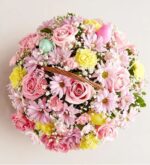 A top-down view of a lush, round floral arrangement in a wicker basket, featuring pink roses, daisies, yellow carnations, and pastel-colored Easter eggs.