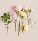 A minimalist flat lay arrangement of various spring flowers and a single yellow egg on a pale pink background. The collection includes a pink rose, carnations, daisies, baby's breath, and a leafy green stem.