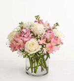 A round glass vase holding a bouquet of blush pink and white flowers, including roses, alstroemeria, stock, and baby’s breath, set against a clean white background.
