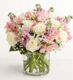 A round glass vase filled with a lush arrangement of ivory white roses, soft pink alstroemeria, pale pink stock, and baby's breath, with green stems visible through the clear water.
