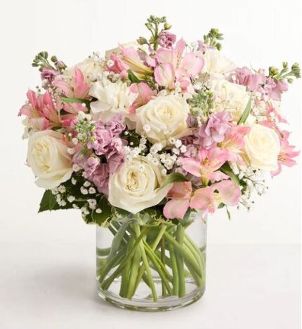 A round glass vase filled with a lush arrangement of ivory white roses, soft pink alstroemeria, pale pink stock, and baby's breath, with green stems visible through the clear water.