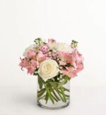 Elegant bouquet of blush pink alstroemeria, ivory roses, and white carnations arranged with greenery in a clear glass cylinder vase, set against a white background.