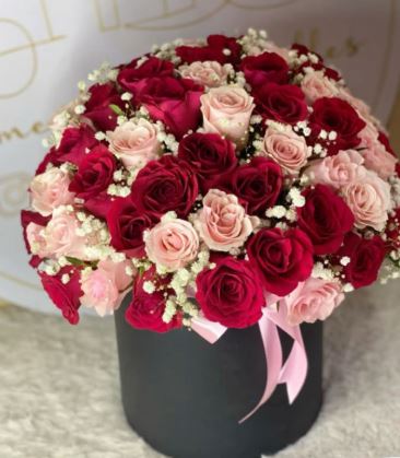 A dense, dome-shaped floral arrangement of deep red and light pink roses mixed with white baby's breath, presented in a cylindrical matte black gift box with a pink satin ribbon.