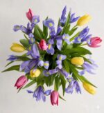 A top-down view of a vibrant spring bouquet featuring purple irises with yellow centers, bright yellow tulips, and pink tulips, all arranged with fresh green leaves against a neutral white background.