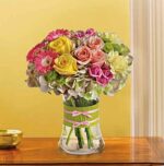 A colorful mixed bouquet in a clear glass vase with lime green ribbon and pink bows. The arrangement features yellow roses, pink spray roses, hot pink gerbera daisies, and pale green hydrangeas, set against a soft yellow wall on a wooden table.