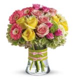 A vibrant mixed flower bouquet in a clear glass vase. The arrangement features hot pink gerbera daisies, yellow and pink roses, pink mini carnations, and light green hydrangeas. The vase is wrapped in a lime green leaf ribbon with two small pink bows.