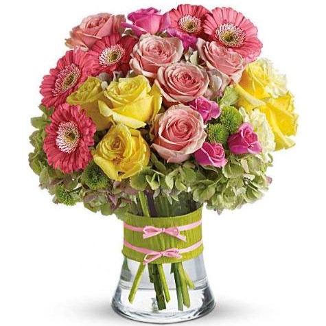 Bright mixed bouquet in a clear glass vase with a green wrap and pink ribbon, featuring pink gerbera daisies, soft pink roses, yellow roses, light green hydrangeas, and small pink accents.