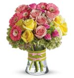 Bright mixed bouquet in a clear glass vase with a green wrap and pink ribbon, featuring pink gerbera daisies, soft pink roses, yellow roses, light green hydrangeas, and small pink accents.