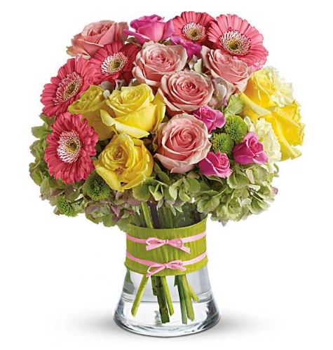 Bright mixed bouquet in a clear glass vase with a green wrap and pink ribbon, featuring pink gerbera daisies, soft pink roses, yellow roses, light green hydrangeas, and small pink accents.