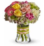 A round mixed flower bouquet in a clear glass vase with a lime green ribbon and two pink bows. The arrangement includes yellow roses, pink roses, pink gerbera daisies, light pink carnations, white hydrangeas, and green button mums.