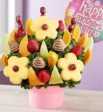 Pink container holding a fruit bouquet with pineapple daisy slices, chocolate-dipped strawberries, red grapes, cantaloupe wedges, and orange slices, arranged over kale. A pink "Happy Mother's Day" balloon is visible in the background.