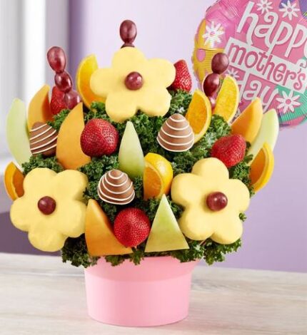 Pink container holding a fruit bouquet with pineapple daisy slices, chocolate-dipped strawberries, red grapes, cantaloupe wedges, and orange slices, arranged over kale. A pink "Happy Mother's Day" balloon is visible in the background.