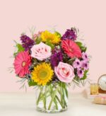 A vibrant mixed flower bouquet in a clear glass cylinder vase, featuring pink roses, yellow sunflowers, bright pink gerbera daisies, purple stock flowers, and lavender chrysanthemums with green foliage against a light pink background.