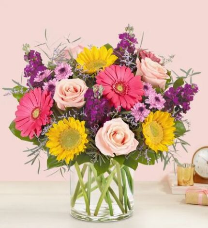 A vibrant floral arrangement in a clear glass cylinder vase featuring pink and yellow gerbera daisies, light pink roses, purple stock flowers, and various green foliage against a soft pink background.