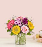 A vibrant mixed flower bouquet in a clear glass cylinder vase, featuring yellow sunflowers, pink roses, hot pink gerbera daisies, purple stock flowers, and light purple pompons with green foliage.