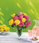 Vibrant bouquet of pink and yellow roses in a clear glass vase on a white outdoor table, with lemons, pink lemonade, and macarons in the background against a bright green backdrop.