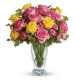 A clear glass vase filled with a lush bouquet of pink and yellow roses, accented with green foliage, set against a white background.