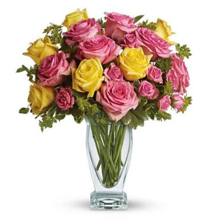 A clear glass vase filled with a lush bouquet of pink and yellow roses, accented with green foliage, set against a white background.