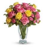 A vibrant bouquet of pink and yellow roses arranged with green foliage in a clear glass vase, set against a white background.