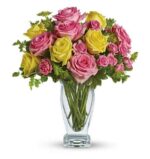 Bouquet of pink and yellow roses with green foliage arranged in a clear glass vase, set against a white background.