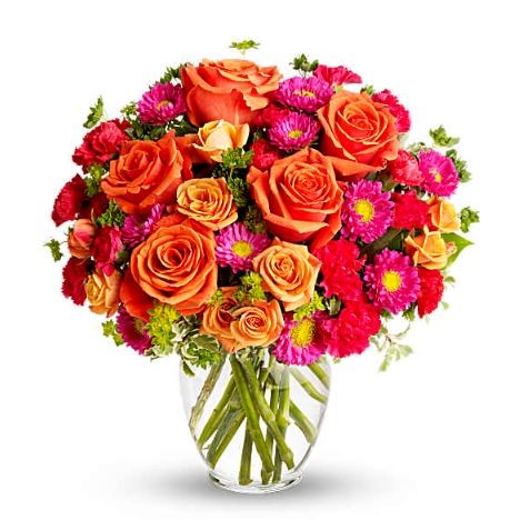 A vibrant bouquet of orange roses, peach spray roses, hot pink carnations, and magenta daisies with yellow centers, arranged with small green accents in a clear glass vase.