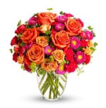 A vibrant bouquet of orange roses, peach spray roses, hot pink carnations, and magenta daisies with yellow centers, arranged with small green accents in a clear glass vase.