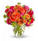 A vibrant floral arrangement in a clear glass vase featuring orange roses, hot pink carnations, magenta chrysanthemums with yellow centers, and green hypericum berries. Stems are visible in the water.