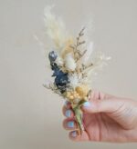 A hand with light blue nail polish holding a small ivory dried flower boutonnière with pampas grass, bleached foliage, and navy accents, wrapped in green twine.