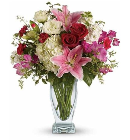 Elegant mixed flower bouquet in a clear glass vase featuring pink stargazer lilies, red roses, white hydrangeas, pink orchids, white lisianthus, and red mini carnations with lush green foliage.