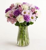 A clear glass vase holding a full bouquet of mixed flowers in shades of lavender, purple, and white. The arrangement includes white roses, purple carnations, light purple daisies with yellow centers, and striped alstroemeria, with green stems visible through the vase.