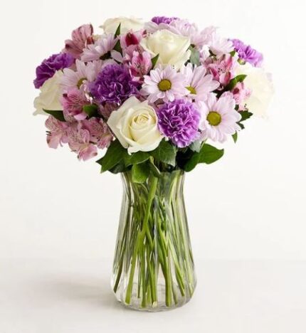 A clear glass vase holding a full bouquet of mixed flowers in shades of lavender, purple, and white. The arrangement includes white roses, purple carnations, light purple daisies with yellow centers, and striped alstroemeria, with green stems visible through the vase.