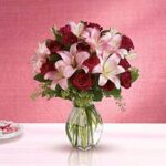 A lush bouquet of deep red roses and soft pink lilies with green foliage in a clear glass vase, set against a textured pink background. A small dish of pink and white candies sits beside it on a white surface.