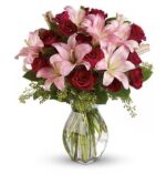 A lush floral arrangement in a clear glass vase, featuring deep red roses, soft pink lilies with speckled petals, and small green filler flowers.