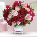 A lush bouquet of red, pink, and fuchsia roses in a square glass vase, arranged in a full, rounded shape against a soft pink background.
