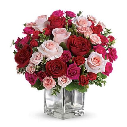 A lush round bouquet of red, pink, and hot pink roses with small green accents, arranged in a clear square glass vase on a white background.