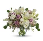 Elegant floral arrangement in a clear glass vase featuring white lilies, lavender roses, white carnations, and stock, accented with eucalyptus and delicate pink filler flowers.