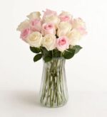 A bouquet of two dozen light pink and cream roses arranged in a clear, fluted glass vase against a white background.