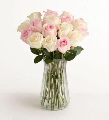 A bouquet of two dozen light pink and cream roses arranged in a clear, fluted glass vase against a white background.