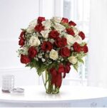 Red and white long-stemmed roses arranged in a clear glass vase with baby's breath and greenery, tied with a large red satin ribbon, displayed on a white table by a bright window.