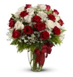 Elegant glass vase filled with long-stemmed red and white roses, accented with baby's breath and lush green ferns, tied with a large red satin bow.