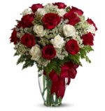 A large floral arrangement in a clear glass vase with a red ribbon bow. The bouquet features a mix of long-stemmed red and white roses, accented with delicate white baby’s breath and lush green fern foliage.